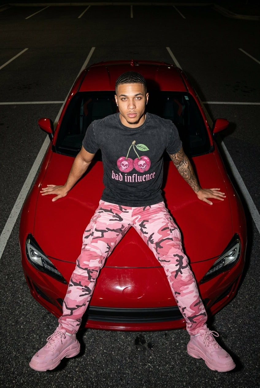 Person wearing a black t-shirt with cherry design and pink camouflage pants standing in front of a red car.