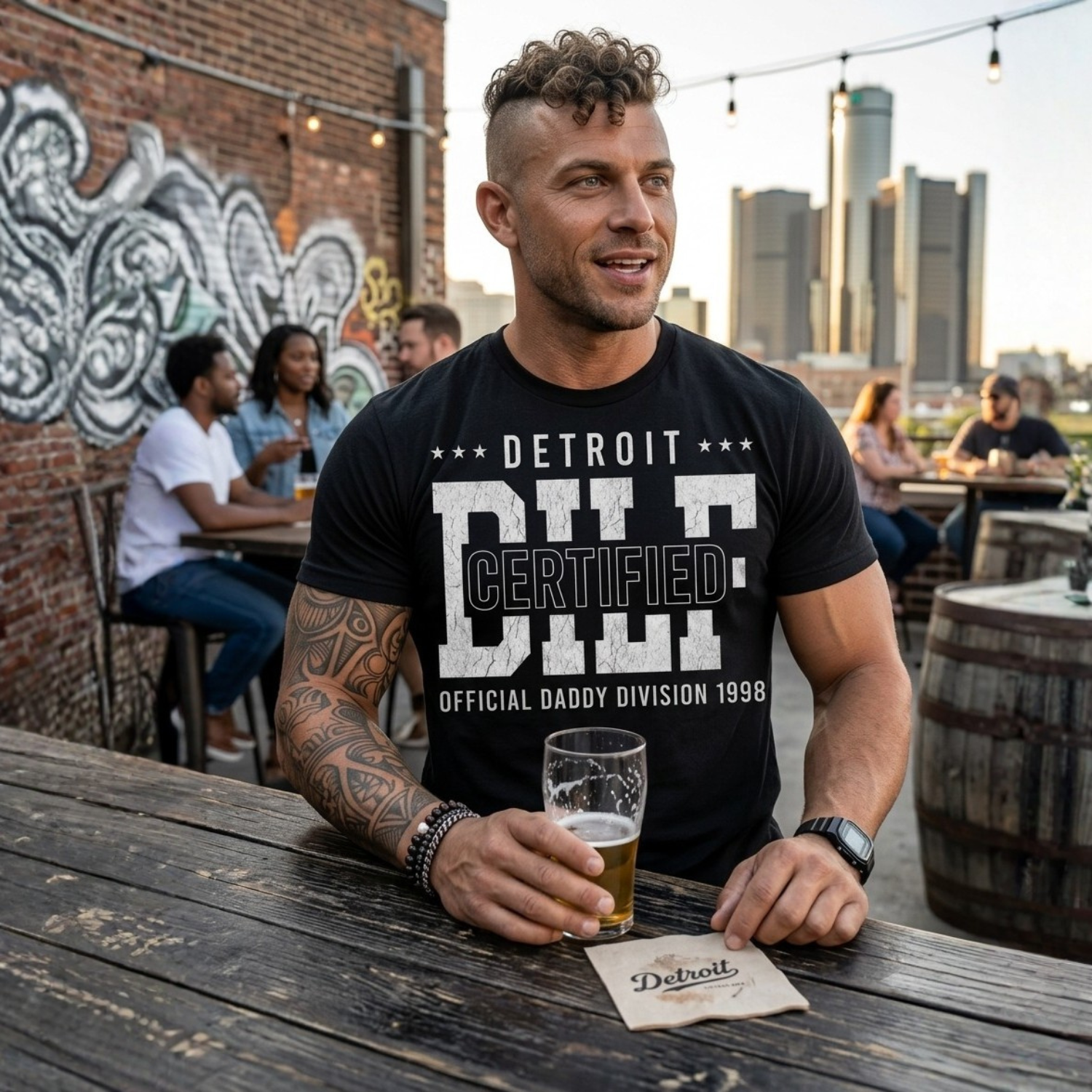 Man holding a beer at an outdoor bar with a city skyline in the background wearing a daddy graphic tee