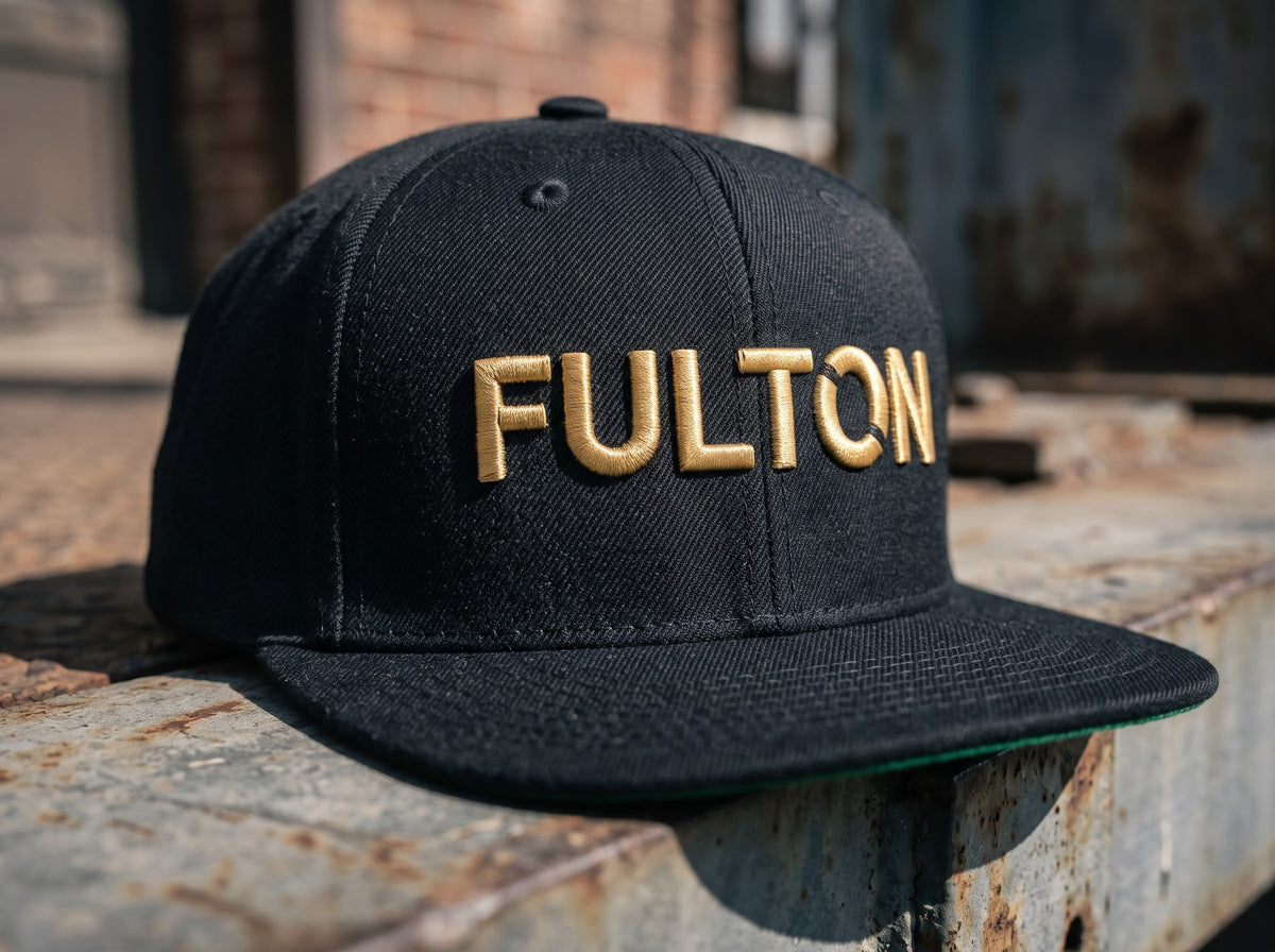 Black flat-brim snapback with raised gold FULTON lettering on a weathered metal ledge.