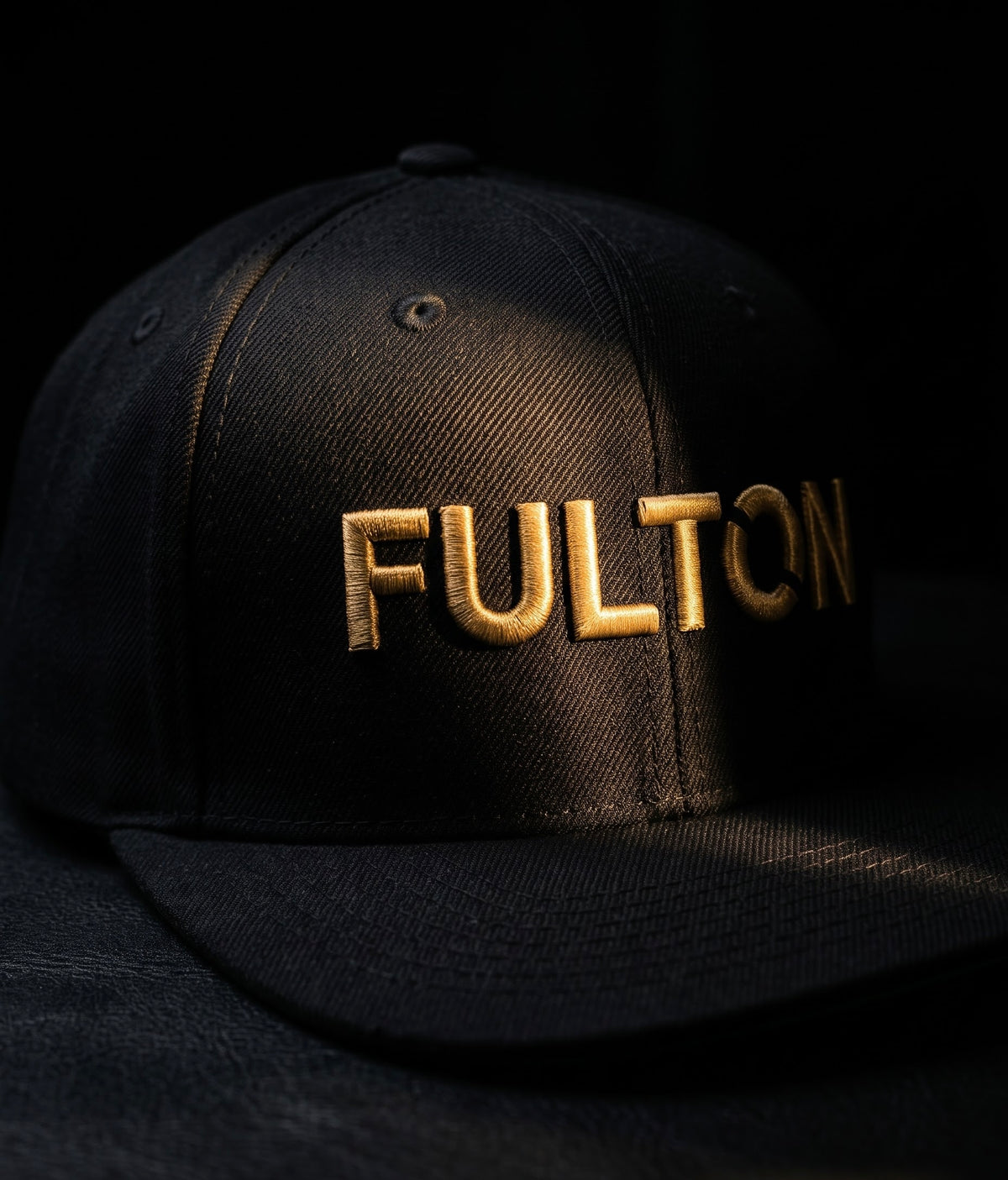 Black flat-brim snapback with raised gold FULTON embroidery against a dark background.