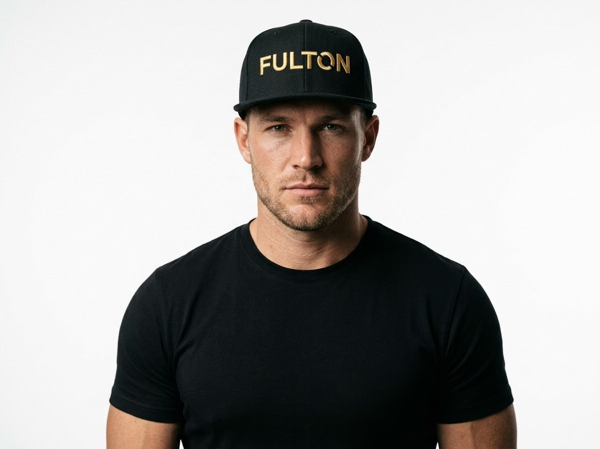 Front portrait of a man wearing a black snapback with gold FULTON lettering and a black T-shirt against a white background.