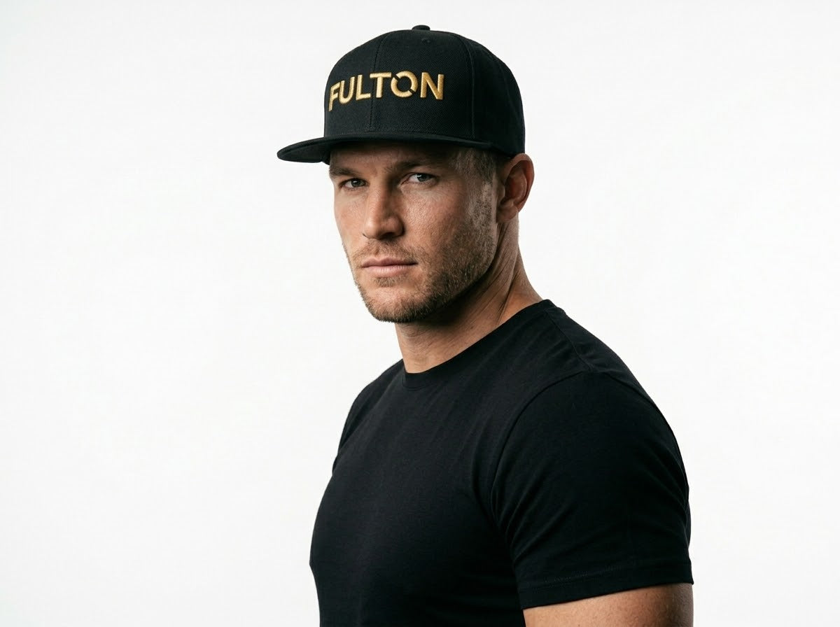 Three-quarter portrait of a man wearing a black snapback with gold FULTON lettering against a white background.